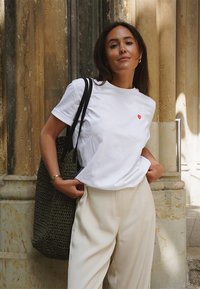 Weißes Baumwoll-T-Shirt mit einem kleinen roten Herzgrafik, kombiniert mit cremefarbenen weit geschnittenen Hosen und einer strukturierten grünen Einkaufstasche.