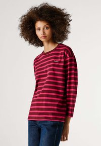 Dugotrajna majica s prugama u boji tamnocrvene i ružičaste, s okruglim izrezom i izvezenim logotipom na prsima, uparena s plavim trapericama.