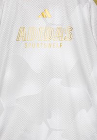 Hvit atletisk skjorte med teksturert stoff, som har et subtilt mønster, gull "ADIDAS"-logo og teksten "SPORTSWEAR" under den i gull.