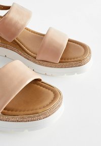 Light pink sandals feature two wide straps, a cushioned beige footbed, and a white sole with a decorative rose gold embellishment around the edge.