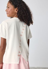Tricou crem cu mâneci scurte, prevăzut cu un imprimeu vertical "TED BAKER" pe cusătura din spate, cu tiv în formă de V și textură moale din bumbac.