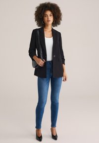 Černý blazer s jedním knoflíkem, v kombinaci s bílým topem a modrými skinny džínami. Drží černou kabelku přes rameno a má na sobě černé špičaté podpatky.