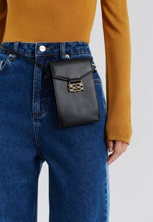 Sort læderpose med gulddetaljer, fastgjort til en blå denimlomme, stylet med en karrygul bluse med lange ærmer.