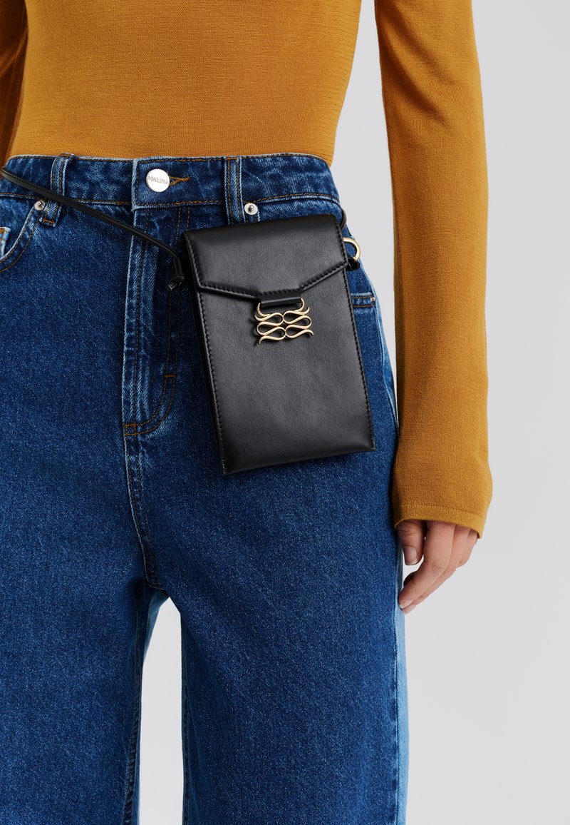 Μαύρη δερμάτινη θήκη με χρυσές μεταλλικές λεπτομέρειες, προσαρτημένη σε μπλε τσεπάκι από τζιν, συνδυασμένη με μουσταρδί μακρυμάνικη μπλούζα.