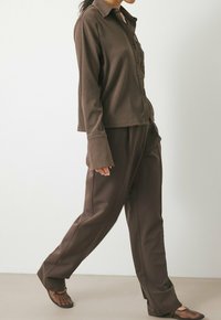Bruine, geribbelde langermige shirt met knopen, gecombineerd met losse, donkerbruine broek en zwarte, minimalistische sandalen. Glad oppervlak overal.