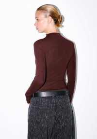 Burgundy top met lange mouwen en een getailleerd ontwerp, gecombineerd met hooggesneden zwarte, gestructureerde broek, voorzien van een zwarte riem en subtiele patronen.