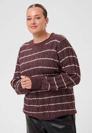 Femme souriante aux cheveux plaqués en arrière, portant un pull en maille bordeaux à rayures horizontales blanches et un pantalon en cuir noir sur un fond gris.