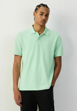 Fiatal férfi fonott hajjal, mentazöld pólóingben és fekete nadrágban, egyszínű világos háttér előtt áll.