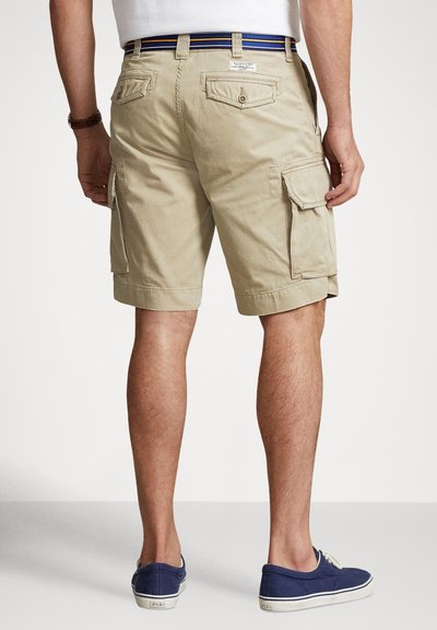 Beżowe szorty cargo z wieloma bocznymi kieszeniami, o prostym kroju i przeszywanym obszyciu. Noszone z niebiesko-białym paskiem w paski i granatowymi butami.