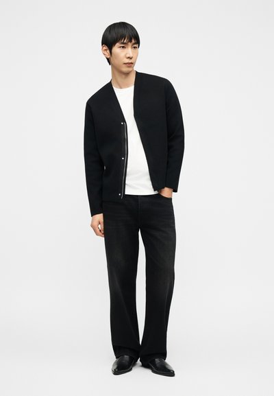 Άνδρας με μαύρο ζακέτα με φερμουάρ, λευκή μπλούζα, φαρδιά μαύρα τζιν και μαύρα παπούτσια, στέκεται με το ένα χέρι στην τσέπη, κοιτάζοντας πλάγια.