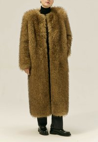 Brązowy płaszcz z sztucznego futra, długi, z szerokim kołnierzem, o fakturowanym, oversize'owym kroju, zapinany z przodu, noszony na czarną golfową bluzkę i ciemne dżinsy. Czarne buty.