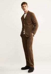 Completo marrone con giacca su misura dotata di taschino a petto e pantaloni coordinati. Indossato con una camicia bianca e scarpe scure.