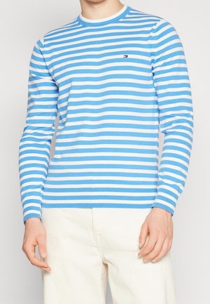 Mann in einem hellblauen und weißen horizontal gestreiften Langarmshirt und cremefarbenen Hosen, der vor einem schlichten Hintergrund steht.