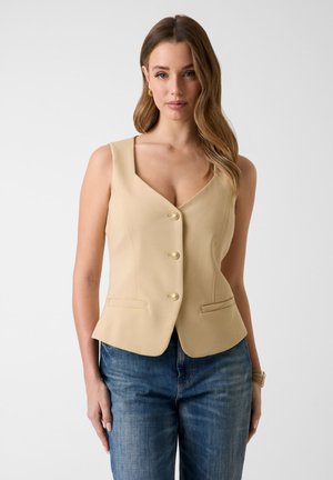 Frau mit langen, welligen Haaren, die goldene Ohrringe, eine beige ärmellose, geknöpfte Weste, blaue Jeans und ein goldenes Armband trägt, vor einem schlichten Hintergrund steht.