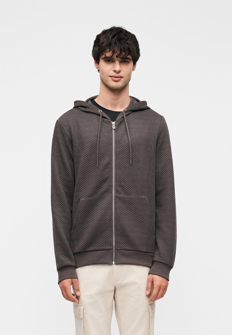 Γκρι φούτερ με φερμουάρ, με υφή σε σχέδιο καπιτονέ, εμπρόσθιες τσέπες και κουκούλα. Συνδυασμένο με ανοιχτόχρωμο παντελόνι.