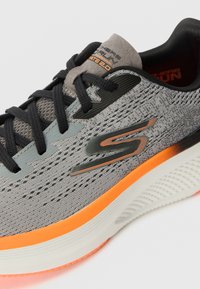 Gray athletic shoe with a breathable mesh upper, black laces, and an orange accent on the sole. Features a textured logo on the side.