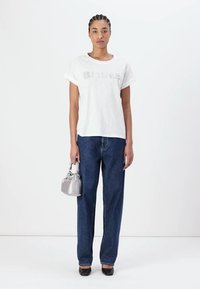 Camiseta blanca de manga corta con el texto "Blauer" en gris, combinada con jeans anchos de tono azul oscuro, sosteniendo un pequeño bolso de mano plateado y zapatos negros.