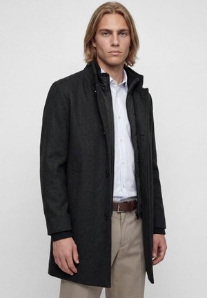 Young man with shoulder-length blonde hair wears a black buttoned coat over a white striped shirt and beige pants against a plain background.