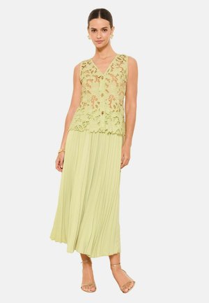 Nő ujjatlan világoszöld csipkés, gombos felsőben, pliszírozott, hozzáillő midi szoknyában és arany nyitott orrú szandálban, oldalán tartott kézzel áll.