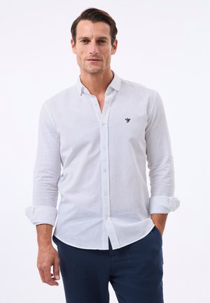 Mand iført hvid skjorte med knapper og opkørte ærmer og navybukser, stående med den ene hånd i lommen mod hvid baggrund.