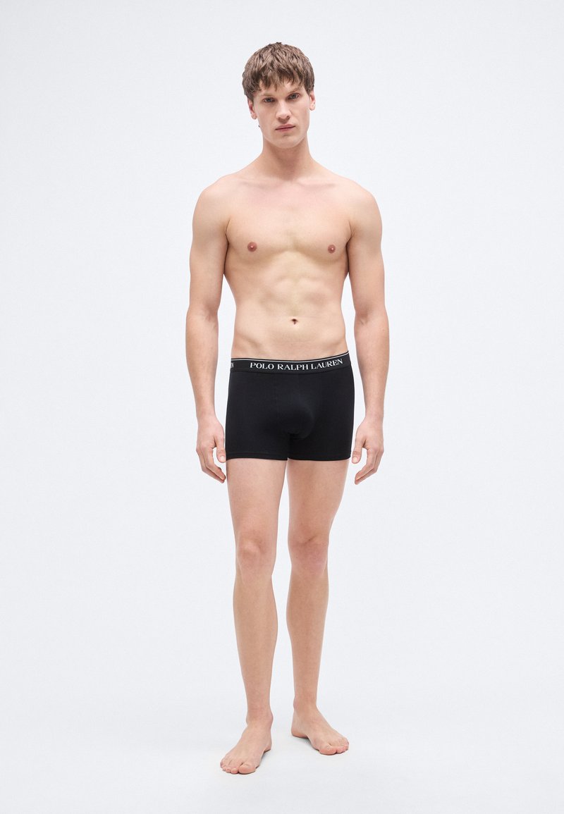 Black boxer briefs made of smooth fabric with a fitted design and a branded waistband. Model stands barefoot against a plain background.