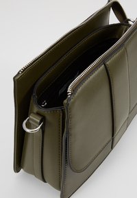 Bolso bandolera rectangular verde oliva con cremallera plateada, bolsillo externo y bordes ribeteados en negro, mostrado abierto para revelar los compartimentos interiores.