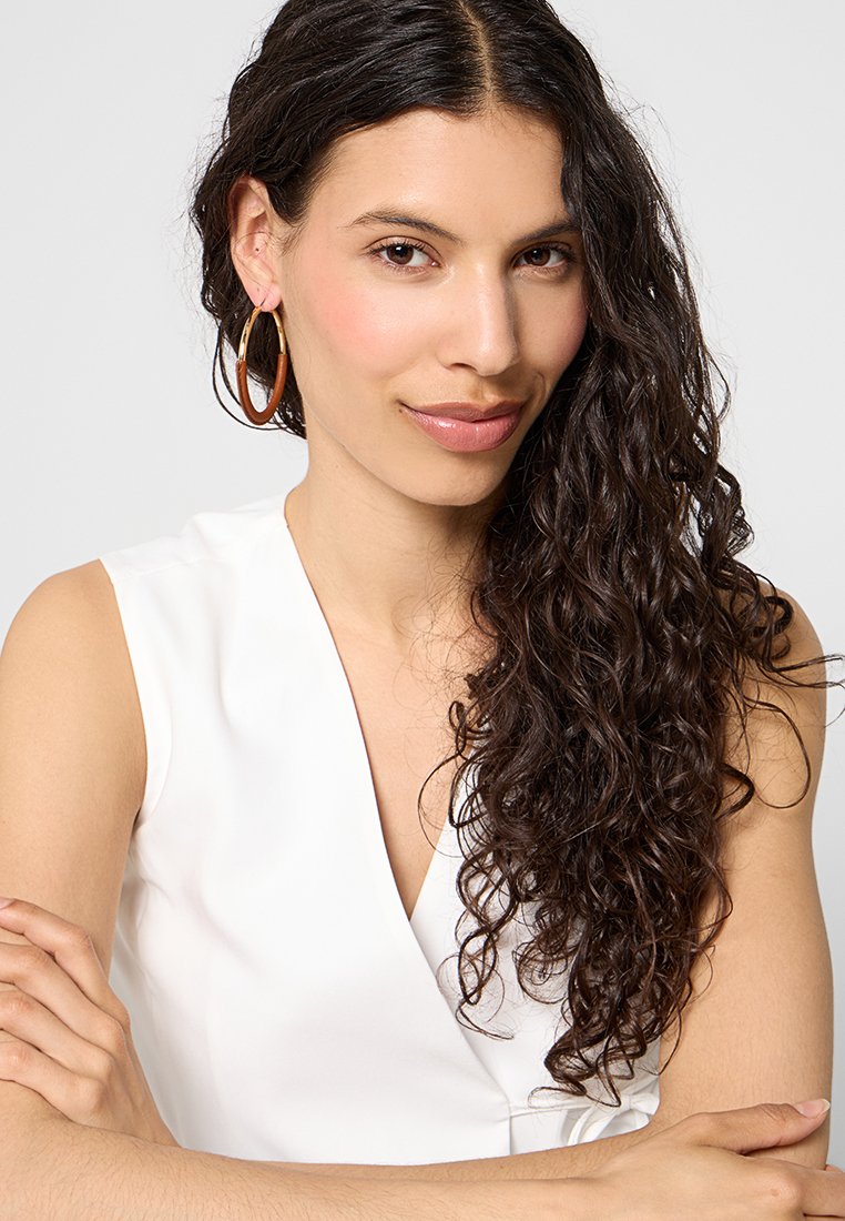 Des cheveux bouclés et foncés entourent un visage avec un maquillage naturel. Vêtue d'un haut blanc sans manches et de grandes boucles d'oreilles rondes en or, les bras croisés.
