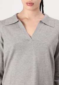 Femme portant un pull à col en V gris clair avec un col replié, laissant apparaître le cou et le haut de la poitrine.