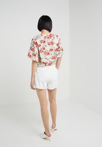 Kukkainen pusero, jossa on lyhyet röyhelöhihat. Siinä on punaisia ja vaaleanpunaisia kukkia vaalealla pohjalla. Yhdistetty valkoisiin shortseihin ja samaa sävyä oleviin avokkaisiin.