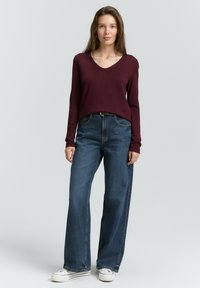 Bordeaux lange mouw V-hals trui gecombineerd met donkerblauwe wijde jeans en witte sneakers met zwarte strepen, staande tegen een lichte achtergrond.