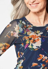 Bluse mit langen Ärmeln in marineblauem Stoff, versehen mit bunten Blumenmustern, transparenter Netzärmel und rundem Ausschnitt. Sichtbare Akzente.