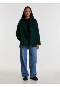 Groen-geruit button-up jack met een ontspannen pasvorm, gecombineerd met blauwe wijde jeans en zwarte chunky laarzen. Soepele textuur, casual ontwerp.