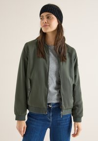 Chaqueta bomber verde con acabado suave, puños acanalados y bolsillos con cremallera, combinada con un suéter gris y jeans de mezclilla azul.