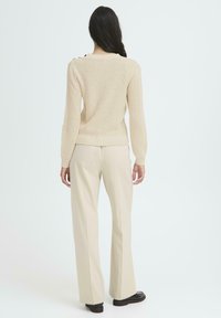 Mujer con cabello largo y oscuro, viste un suéter de punto beige, pantalones beige a juego y mocasines negros, de pie de espaldas sobre un fondo blanco.