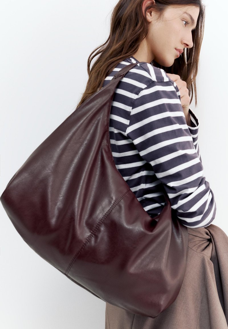 Jeune femme portant une chemise rayée porte un grand sac à bandoulière en cuir marron foncé sur son épaule devant un fond uni.