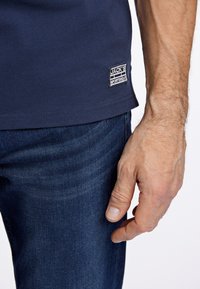 Mörkblå polo med texturerad tyg, med en etikett vid fållen. Bärs tillsammans med mörka denimjeans, handen vilar avslappnat mot byxorna.