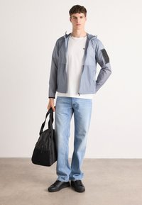 Lightweight, gray zip-up jacket with hood, front pockets, and textured panels; paired with a white t-shirt, light jeans, and black bag.