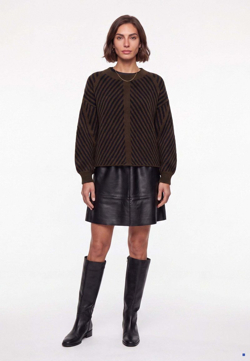 Femme portant un pull rayé marron et noir, une jupe en cuir noire et des bottes noires montantes jusqu'au genou, debout devant un fond blanc.