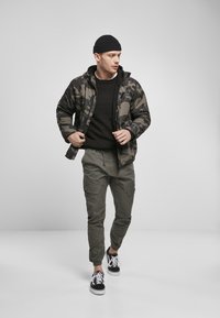 Kamuflasje dunjakke, svart strikkegenser, grønne cargo-bukser og svart-hvite sneakers. Har glidelåslukking og lommer.
