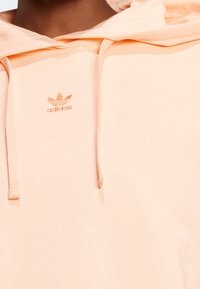 Persikofärgad Adidas-huvtröja med texturerad tyg, dragsko och en liten präglad logotyp på bröstet. Mjuk och lätt design.