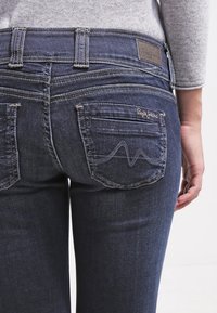 Jeans de mezclilla azul oscuro con un diseño ajustado, que cuentan con bolsillos traseros con costuras onduladas y una etiqueta de cuero cerca de la cintura.