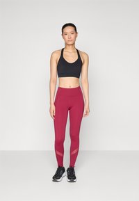 Soutien-gorge de sport noir, leggings taille haute bordeaux avec des accents en panneaux transparents. Modèle portant des chaussures de sport noires, se tenant contre un arrière-plan uni.