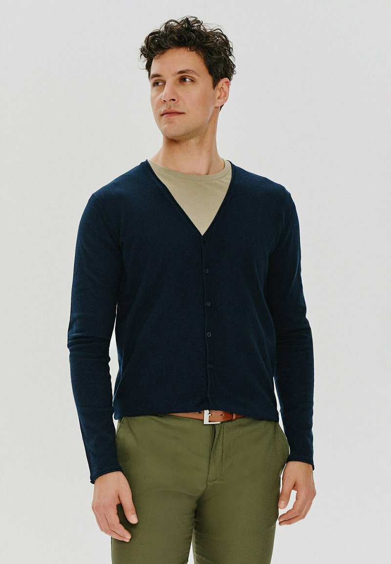 Granatowy kardigan z dekoltem w serek, długimi rękawami i zapięciem na guziki, noszony na beżowej koszuli, w zestawieniu z oliwkowymi spodniami.