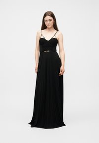 Νεαρή γυναίκα με μακριά καστανά μαλλιά, ντυμένη με μια μαύρη φόρεμα μέχρι το πάτωμα, με λεπτές τιράντες και εφαρμοστό κορσέ, που στέκεται μπροστά σε απλό λευκό φόντο.