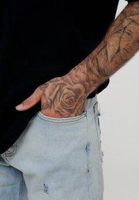Lyseblå slitte jeans med en synlig lomme. En hånd med en rosetatovering hviler på lommen. Skjorten er svart og litt løs i passformen.