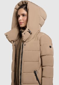 Béžová prošívaná bunda s vysokým límcem a velkou kapucí. Má černé zipy a knoflíky. Hladká textura s prošívaným vzorem.