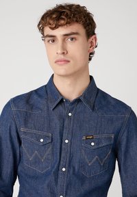 Denim overhemd in donkerblauw met een puntkraag, twee borstzakken met zigzagstiksel en witte drukknoopjes. Wrangler-logo op de zak.