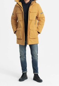 Parka aislante amarilla con múltiples bolsillos frontales, un bolsillo con cremallera en el pecho y una capucha desmontable, combinada con unos vaqueros oscuros y botas negras.