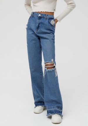 Person wearing high-waisted, loose-fit blue jeans with distressed details on thigh and knee, paired with white sneakers and a cropped white sweater.