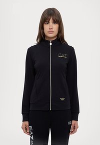 Zwarte ritsjack gemaakt van gladde stof, met een hoge kraag, gouden accenten en logodetails op de borst en zoom.